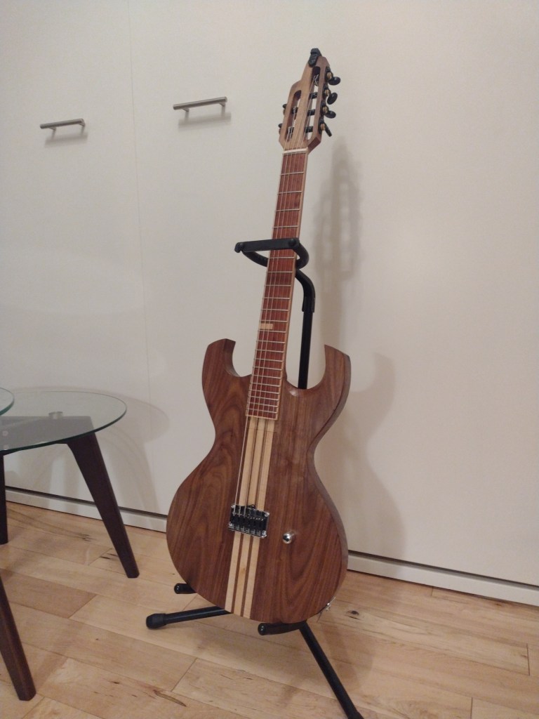 A guitar sits in a black guitar stand. The guitar has a natural finish over walnut, maple and cherry. It has a slotted headstock and no visible pick-ups.