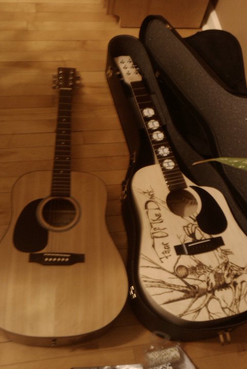 Two guitars lay side by side. A left handed dreadnaught style guitar is on the left. On the right the guitar I built, dreadnaught style with a cut away. The Iron Maiden, Fear of the Dark, album art is wood burned into the top and poker chips are inlayed into the fretboard.