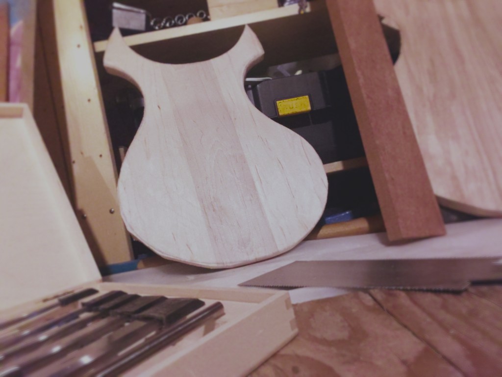 The image focuses on a 3 piece maple top with the centre piece darker in tone than the outer pieces. A red cherry neck blank and ash wood back sit next to it. The foreground is filled with slightly out-of-focus chisel set and Japanese saw.