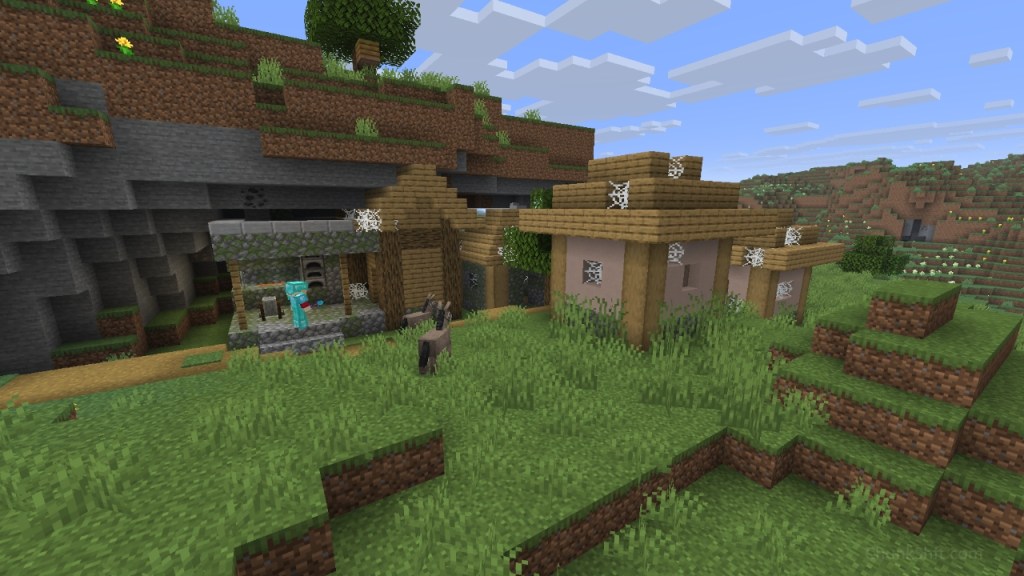 An abandoned Village cuts into the wall of a small cliff. The player, covered in diamond armour can be seen entering the run down blacksmiths forge. 